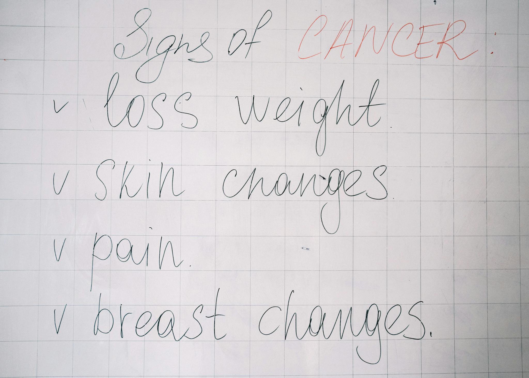 List of cancer symptoms handwritten on a whiteboard including weight loss and skin changes.